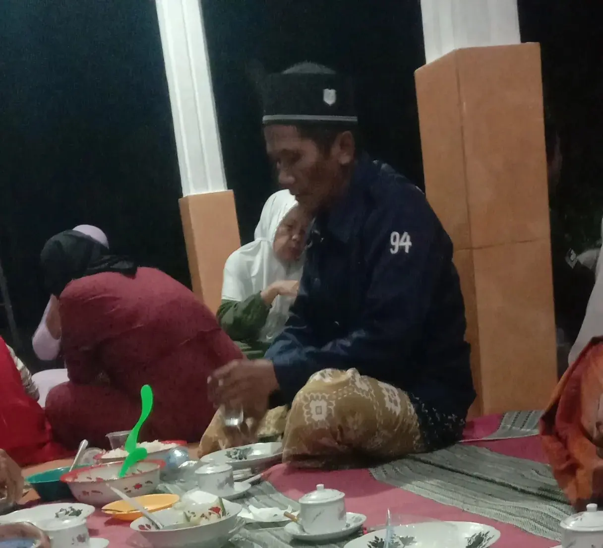 Pemilik motor baru sedang melakukan prosesi selamatan sepeda motor dengan membawa nasi tumpeng dan menghias kendaraan dengan janur di halaman rumahnya. Tradisi ini sebagai wujud syukur dan doa keselamatan."